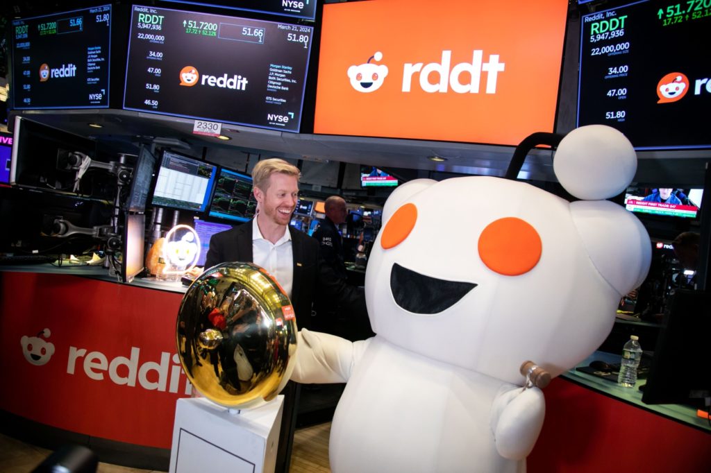 Homem sorridente e mascote Reddit celebram IPO na NYSE, com telas da bolsa e sino dourado em destaque.