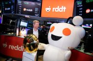Homem sorridente e mascote Reddit celebram IPO na NYSE, com telas da bolsa e sino dourado em destaque.