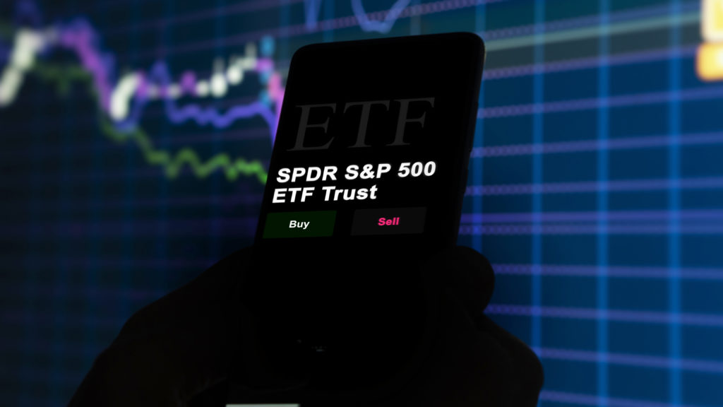 Mão segura celular exibindo tela de negociação de ETF com opções "Comprar" e "Vender". Fundo borrado de gráfico financeiro.
