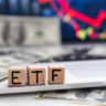 ETF em cubos de madeira, dólares sobre laptop. Tela com gráficos financeiros.