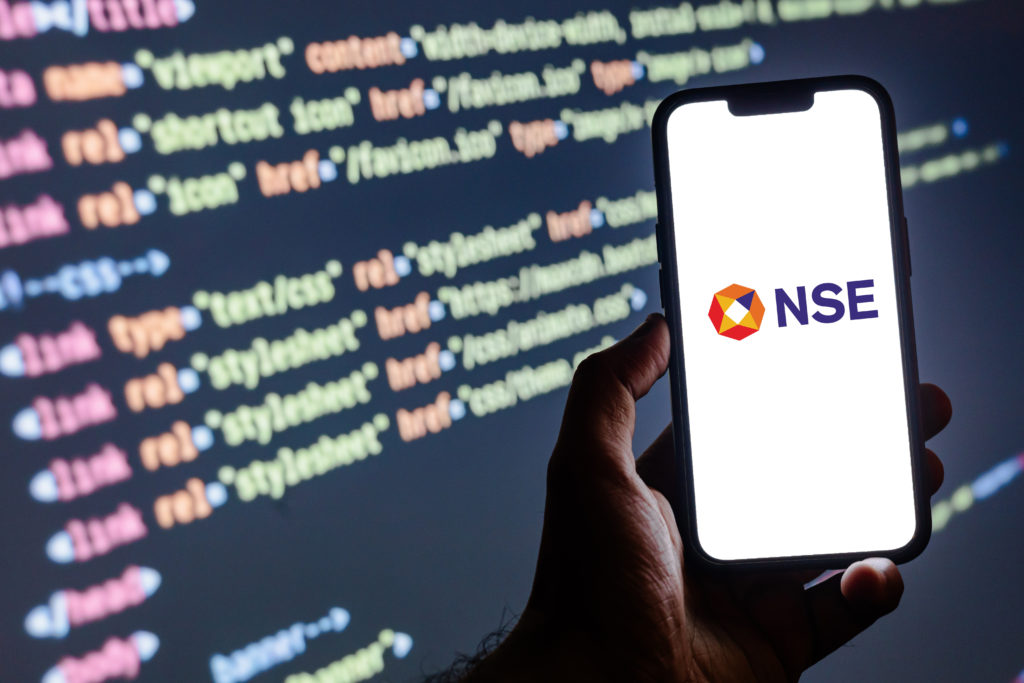 Mão segura smartphone exibindo o logo da NSE, com código de programação digital em segundo plano.