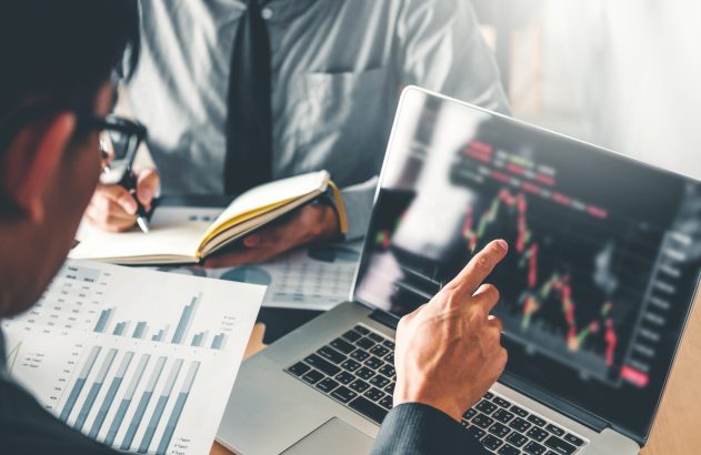 Pessoas analisam gráficos financeiros em laptop e documentos; um aponta para a tela, outro anota.