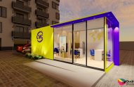 Quiosque modular CVC amarelo e roxo com janelas de vidro, em área externa pavimentada com edifício e carro.