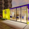 Quiosque modular CVC amarelo e roxo com janelas de vidro, em área externa pavimentada com edifício e carro.