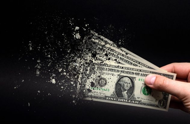 Mão segura notas de dólar que se dissolvem em partículas no ar contra um fundo preto.