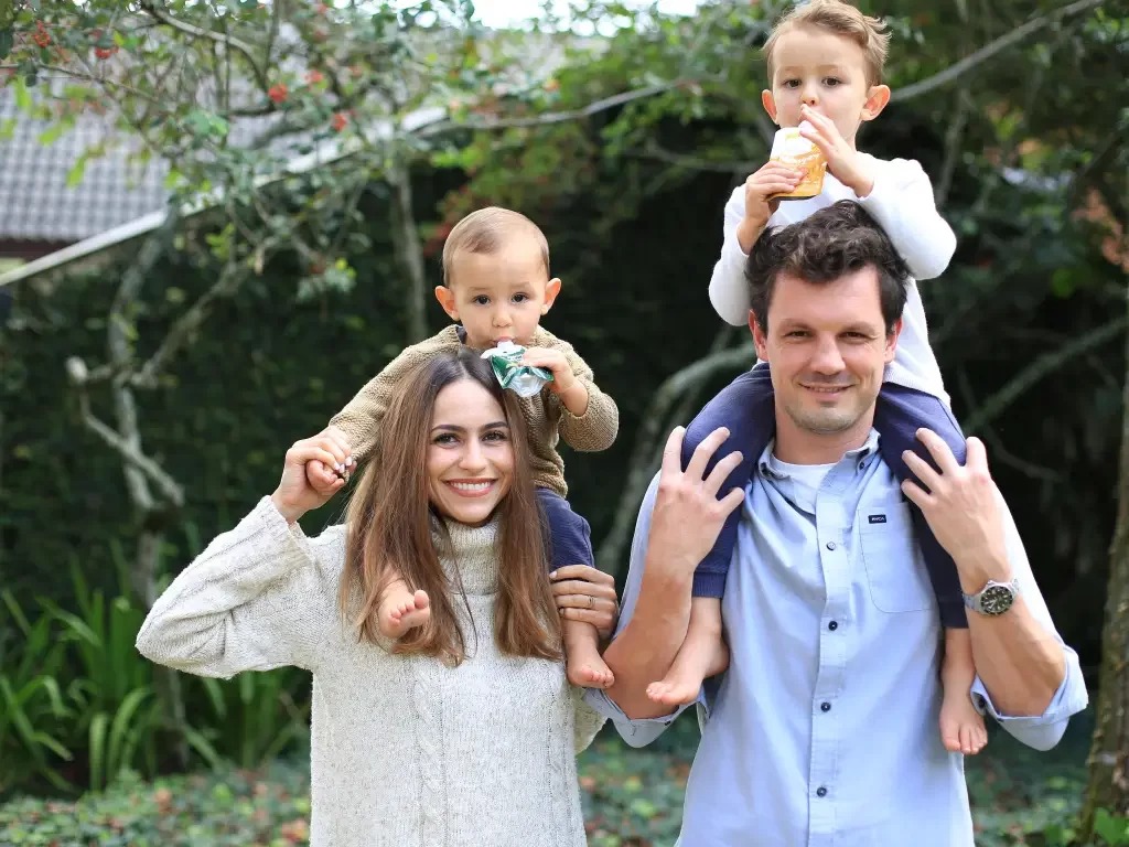 Família de 4: adultos carregam crianças nos ombros, sorrindo ao ar livre.