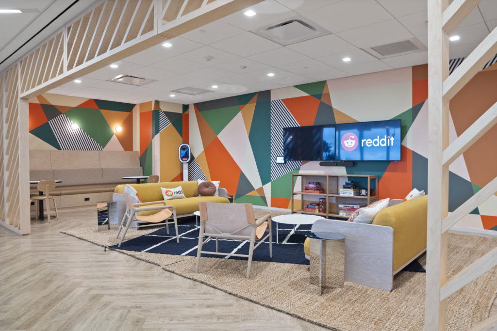 Lounge moderno com mural geométrico colorido, sofás amarelos e TVs exibindo o logo Reddit.