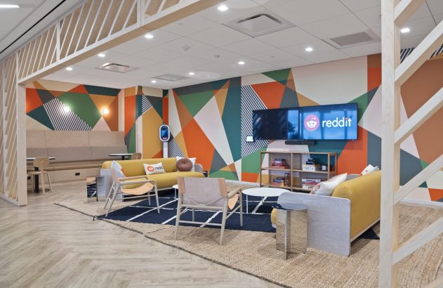 Lounge moderno com mural geométrico colorido, sofás amarelos e TVs exibindo o logo Reddit.
