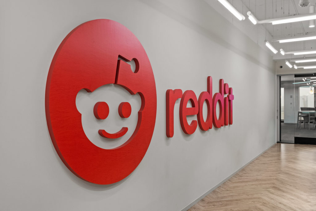 Logo 3D vermelho do Reddit (Snoo e nome) em uma parede branca de escritório, com piso de madeira e iluminação.