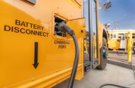 Um ônibus escolar elétrico amarelo sendo carregado. Um cabo de energia está conectado à porta de recarga lateral.
