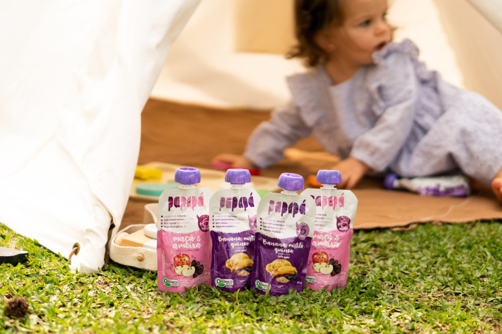 Quatro pouches de papinha Papapá orgânica na grama, com criança e tenda ao fundo. Sabores: maçã/ameixa e banana/mirtilo.