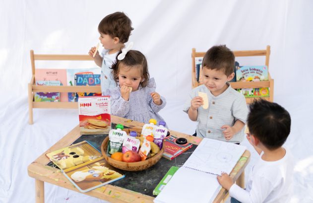 Quatro crianças lancham: três comem biscoitos, uma desenha. Mesa com cesto de frutas e livros. Estantes ao fundo.