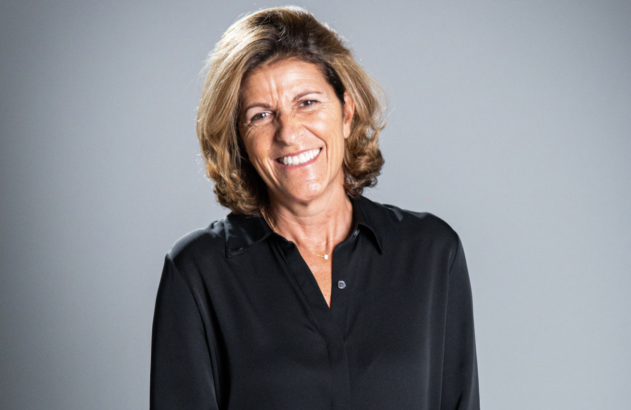 Mulher com cabelo loiro-acastanhado e camisa preta sorrindo para a câmera em fundo cinza.