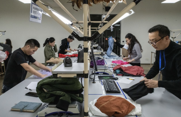 Pessoas inspecionam roupas em mesas com computadores e balanças, em um armazém Shein.