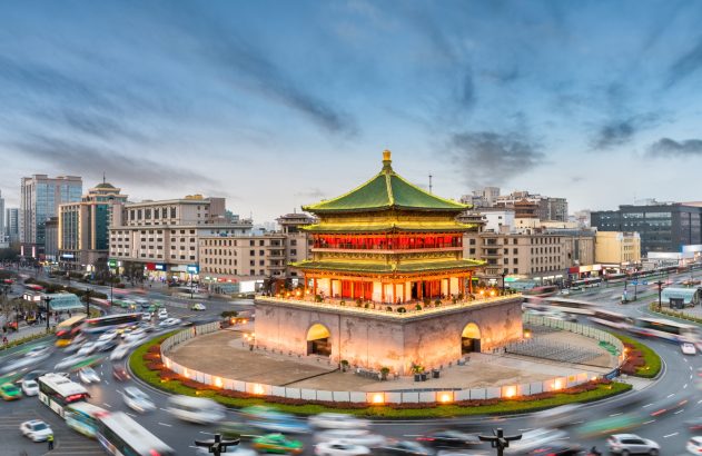 Torre do Sino de Xi'an iluminada, ao centro de rotatória com veículos em tráfego intenso no crepúsculo.