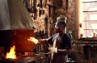 Homem em uma forja, segurando metal quente com faíscas voando. Ele usa óculos e avental.
