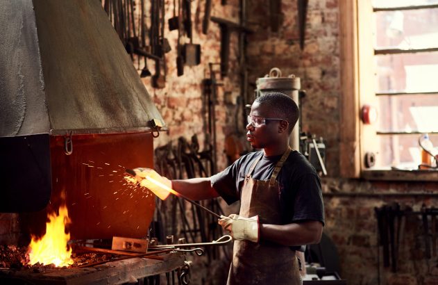 Homem em uma forja, segurando metal quente com faíscas voando. Ele usa óculos e avental.