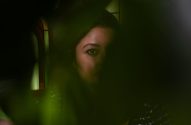 Olho de mulher espreita por entre folhagem verde escura e borrada.