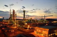 Vista elevada de refinaria em operação ao anoitecer, com torres, tanques e luzes intensas contra o céu crepuscular.