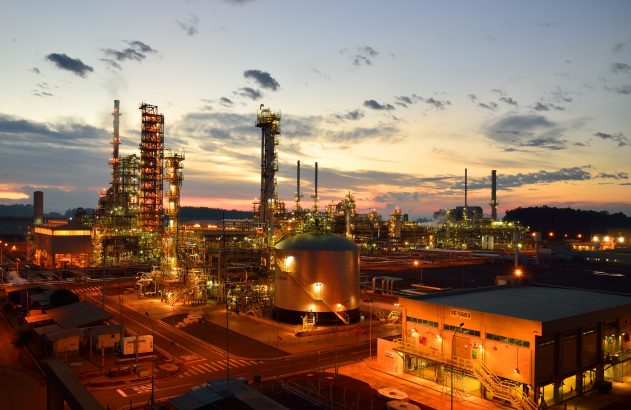 Vista elevada de refinaria em operação ao anoitecer, com torres, tanques e luzes intensas contra o céu crepuscular.