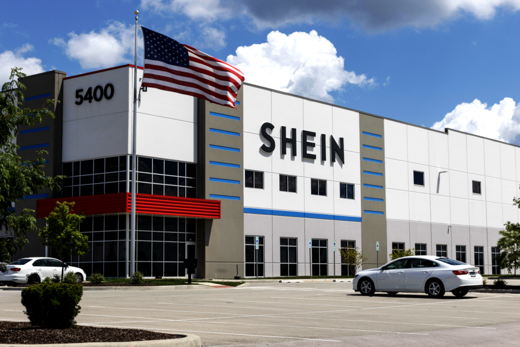 Edifício SHEIN com bandeira dos EUA e carro branco em estacionamento, sob céu azul.