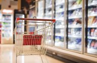 Carrinho de supermercado vazio em corredor com freezers de produtos.