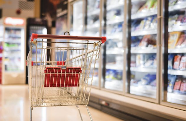 Carrinho de supermercado vazio em corredor com freezers de produtos.