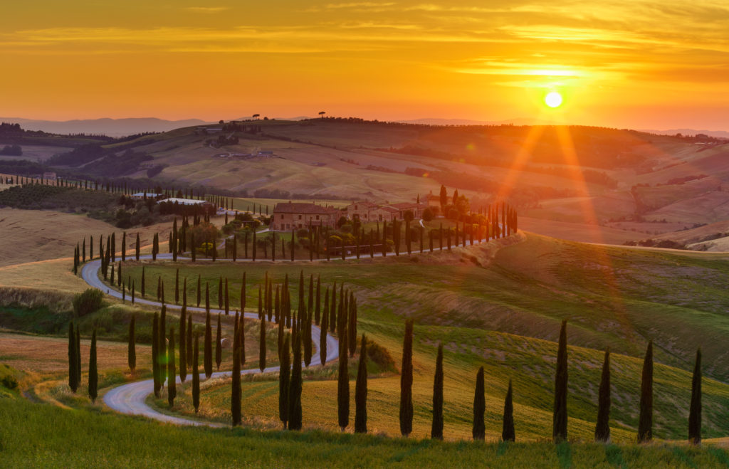 Estrada sinuosa com ciprestes e fazenda na Toscana ao pôr do sol. Sol intenso ilumina as colinas verdes e douradas.