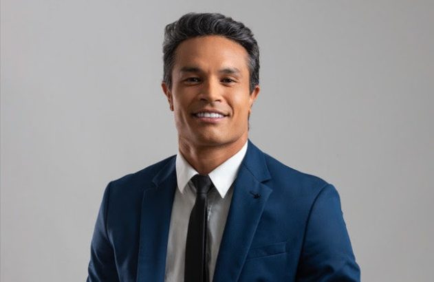 Homem sorridente de terno azul e gravata preta em fundo cinza claro.