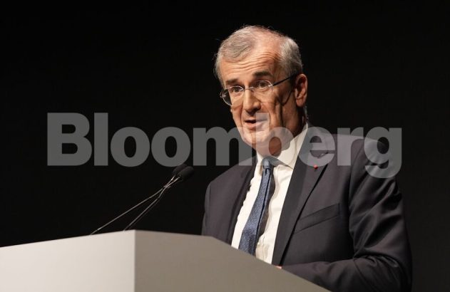 Homem de terno com barba grisalha e óculos fala em pódio com logo Bloomberg ao fundo.