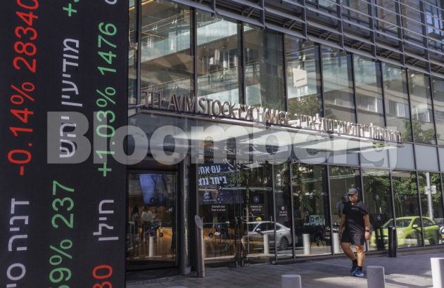 Prédio da Tel Aviv Stock Exchange com painel digital exibindo cotações financeiras em alta e baixa. Um homem caminha.