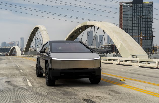 Tesla Cybertruck em ponte de concreto com grandes arcos e skyline urbano. Dia claro.