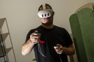 Homem com headset de realidade virtual branco e controles com alça vermelha, jogando.