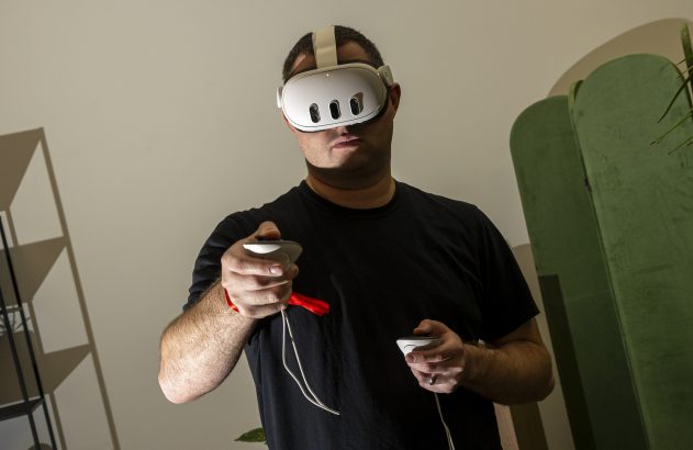 Homem com headset de realidade virtual branco e controles com alça vermelha, jogando.