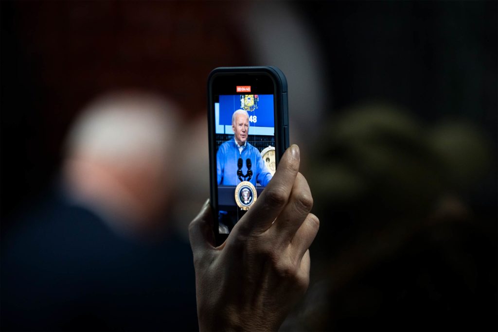 Mão segura smartphone filmando Joe Biden em palanque com selo presidencial.