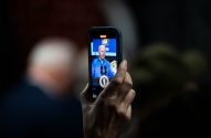 Mão segura smartphone filmando Joe Biden em palanque com selo presidencial.