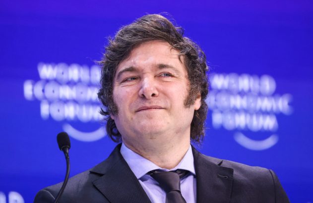 Homem de terno sorrindo e olhando para cima, com microfone. Fundo azul desfocado com 'WORLD ECONOMIC FORUM'.