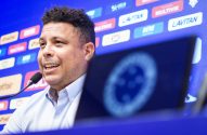 Ronaldo, com camisa azul, fala em microfone em coletiva. Fundo azul com logos, e laptop com logo desfocado à frente.