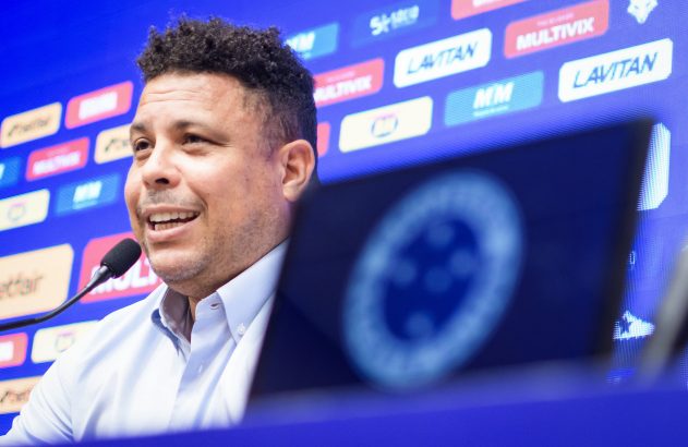 Ronaldo, com camisa azul, fala em microfone em coletiva. Fundo azul com logos, e laptop com logo desfocado à frente.