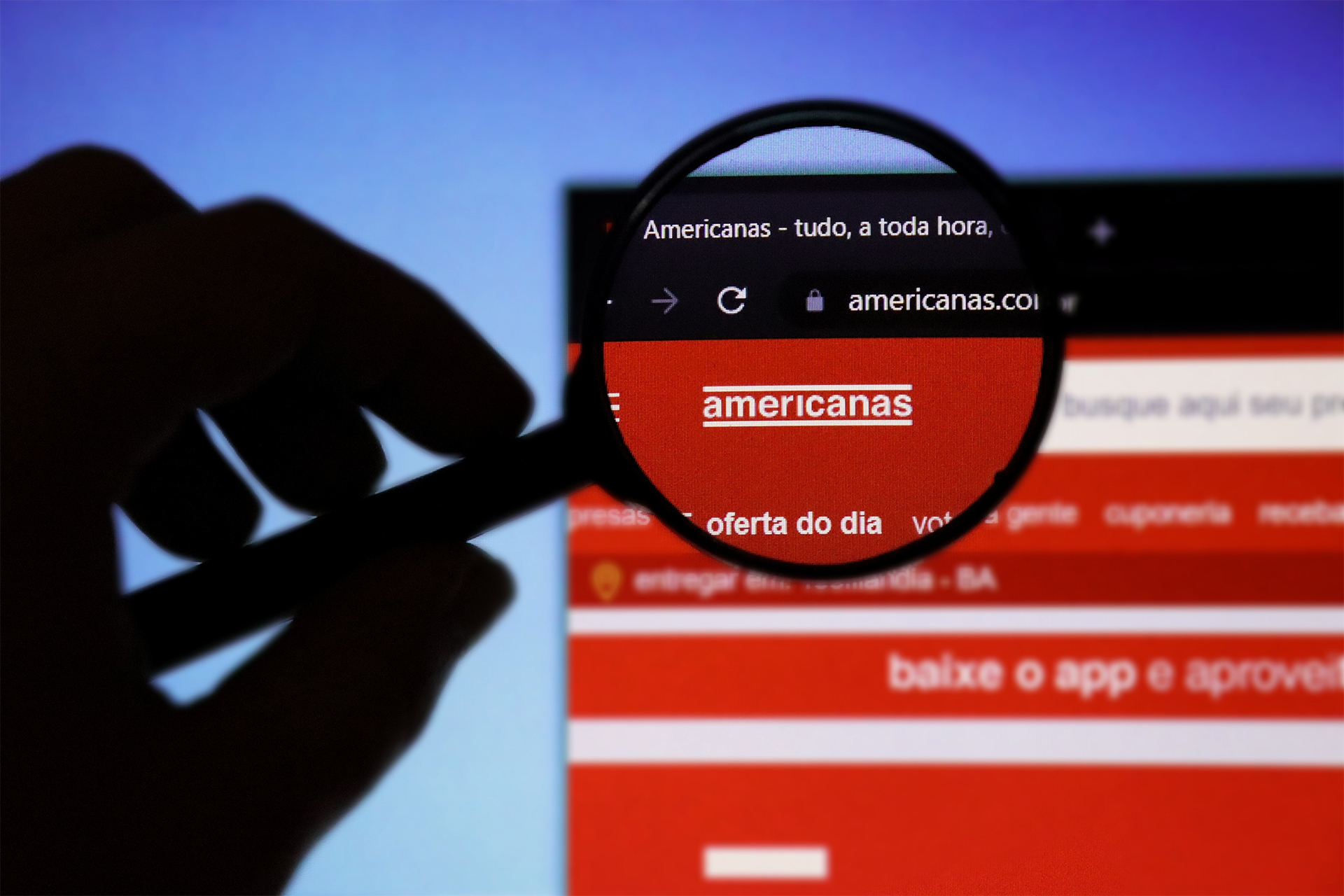 Mão segurando uma lupa sobre o site das Lojas Americanas