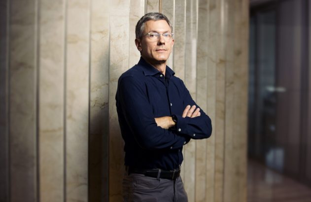 Homem com óculos e camisa azul-marinho, braços cruzados, em pé diante de pilares marmorizados.