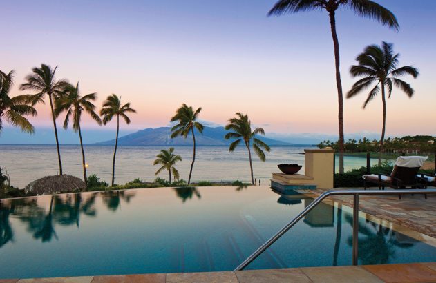 Piscina infinita em paisagem tropical ao pôr do sol, com palmeiras, mar e montanhas no horizonte.