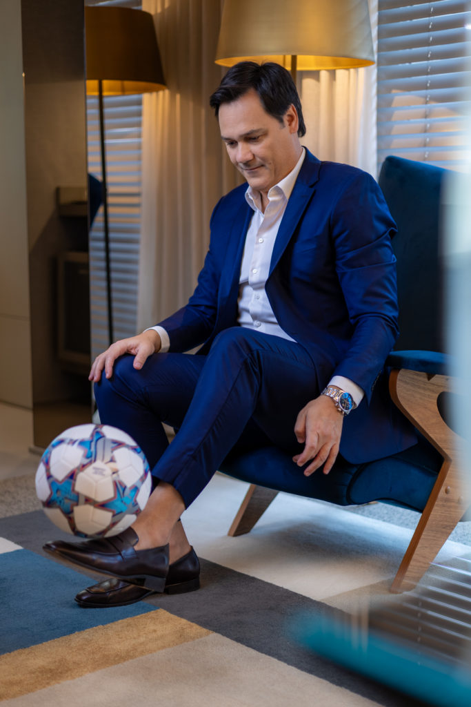 Homem de terno azul sentado em poltrona, equilibrando uma bola de futebol no pé.
