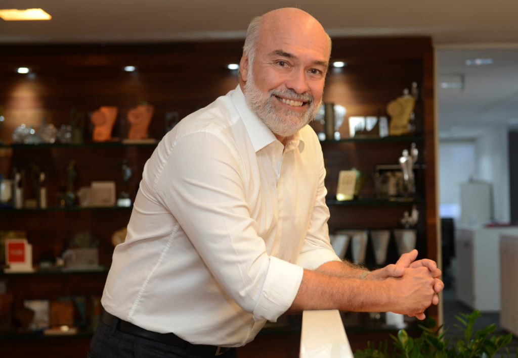 Homem sorridente, calvo e barbudo, de camisa branca, apoiado. Prateleiras com troféus ao fundo.