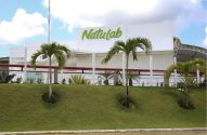 Edifício branco da Natulab com letreiro verde, cercado por um gramado inclinado e palmeiras.