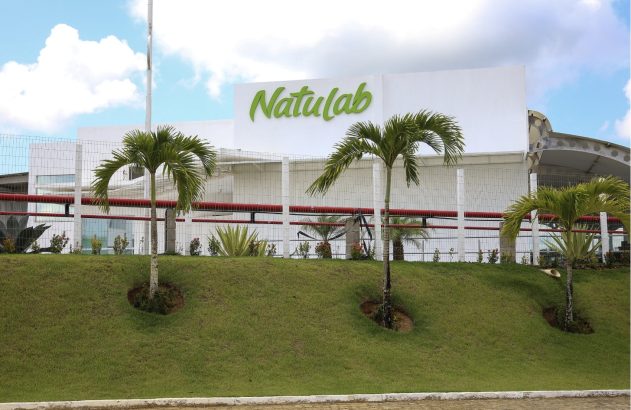 Edifício branco da Natulab com letreiro verde, cercado por um gramado inclinado e palmeiras.