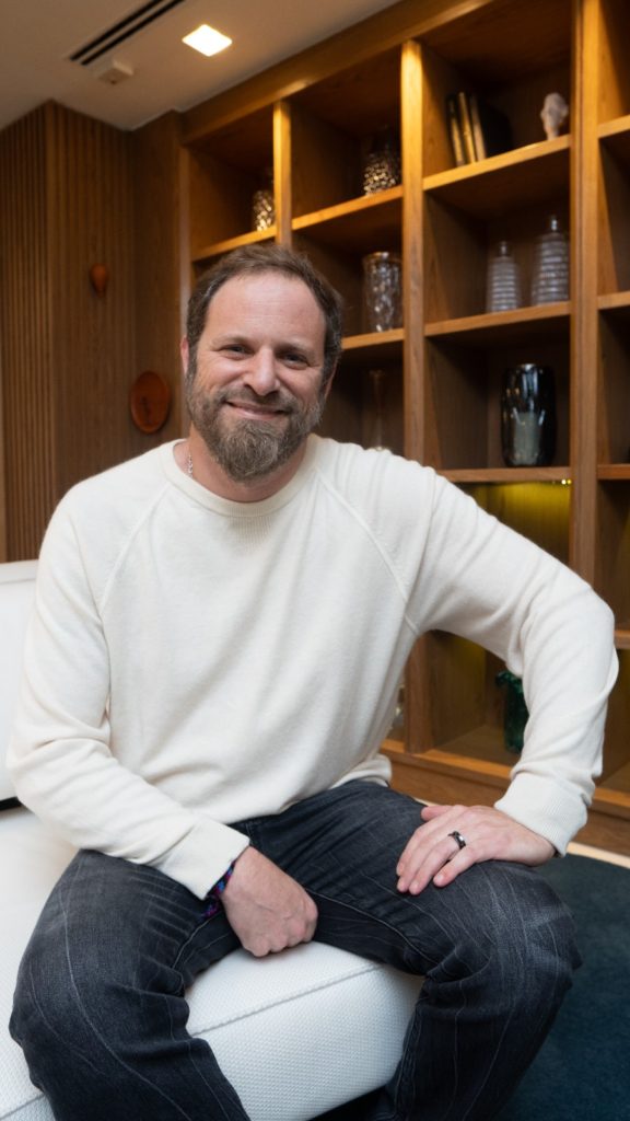 Homem barbudo sorrindo, vestindo suéter claro e jeans, sentado em sofá branco com estante de madeira ao fundo.