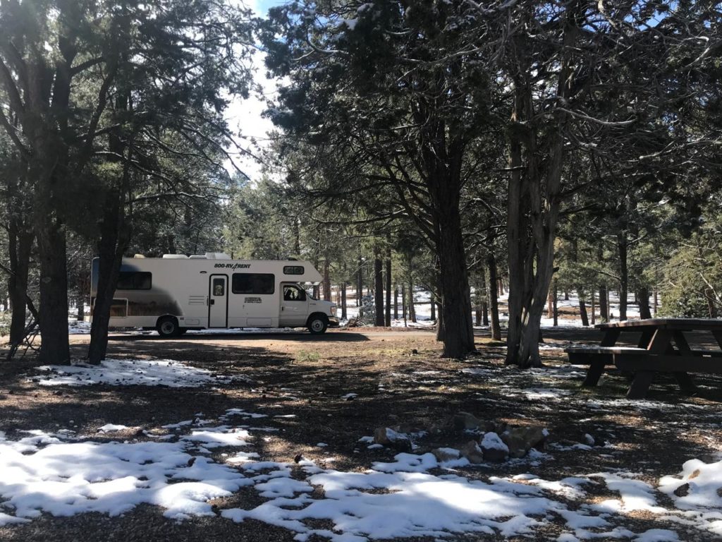 Motorhome branco em acampamento de inverno com neve, árvores, mesa de piquenique e fogueira.