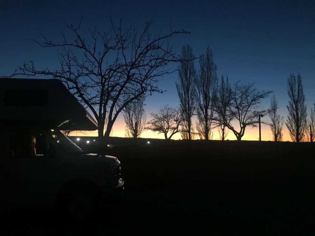 Veículo de campismo e árvores silhuetados ao amanhecer ou entardecer, com faixa de luz no horizonte.