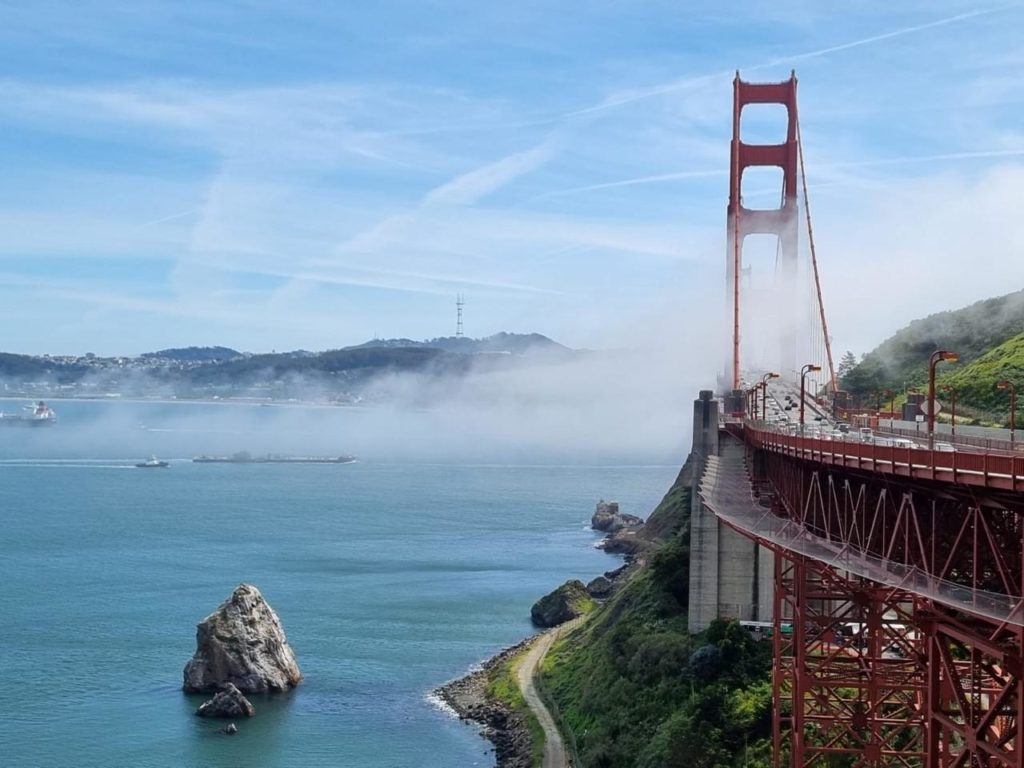 Ponte Golden Gate parcialmente envolta em névoa. Tráfego, barcos na baía e morros distantes.
